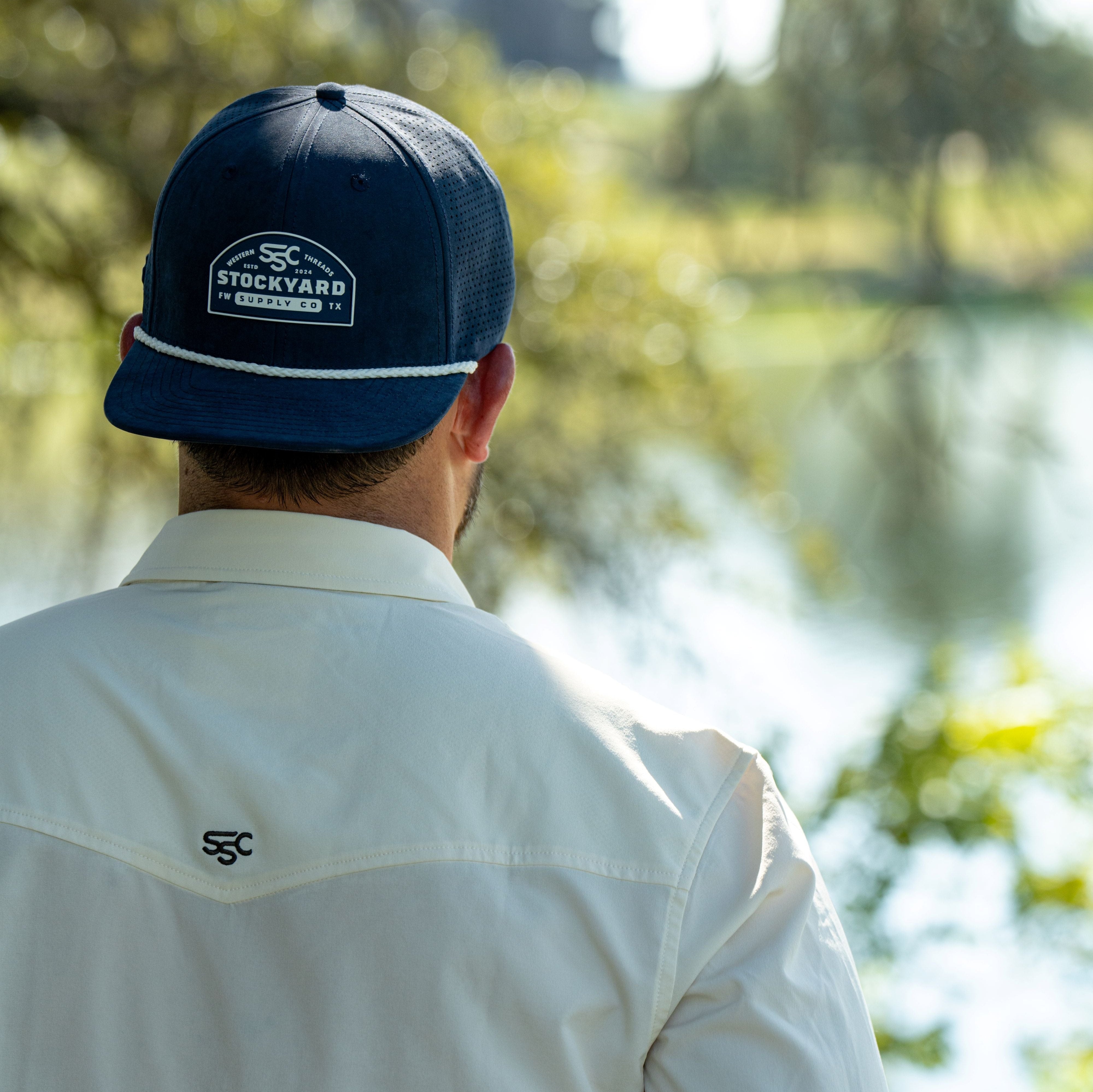 Ryan Dukes wearing the cream colored long sleeve performance pearl snap and a backwards Navy Champion suede hat facing a lake and back towards the camera. 