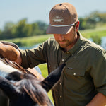 Eric Nelsen wearing the Coyote colored Champion hat and faded olive long sleeve performance pearl snap saddling a horse. 