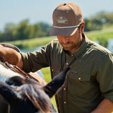 Eric Nelsen wearing the Coyote colored Champion hat and faded olive long sleeve performance pearl snap saddling a horse. 