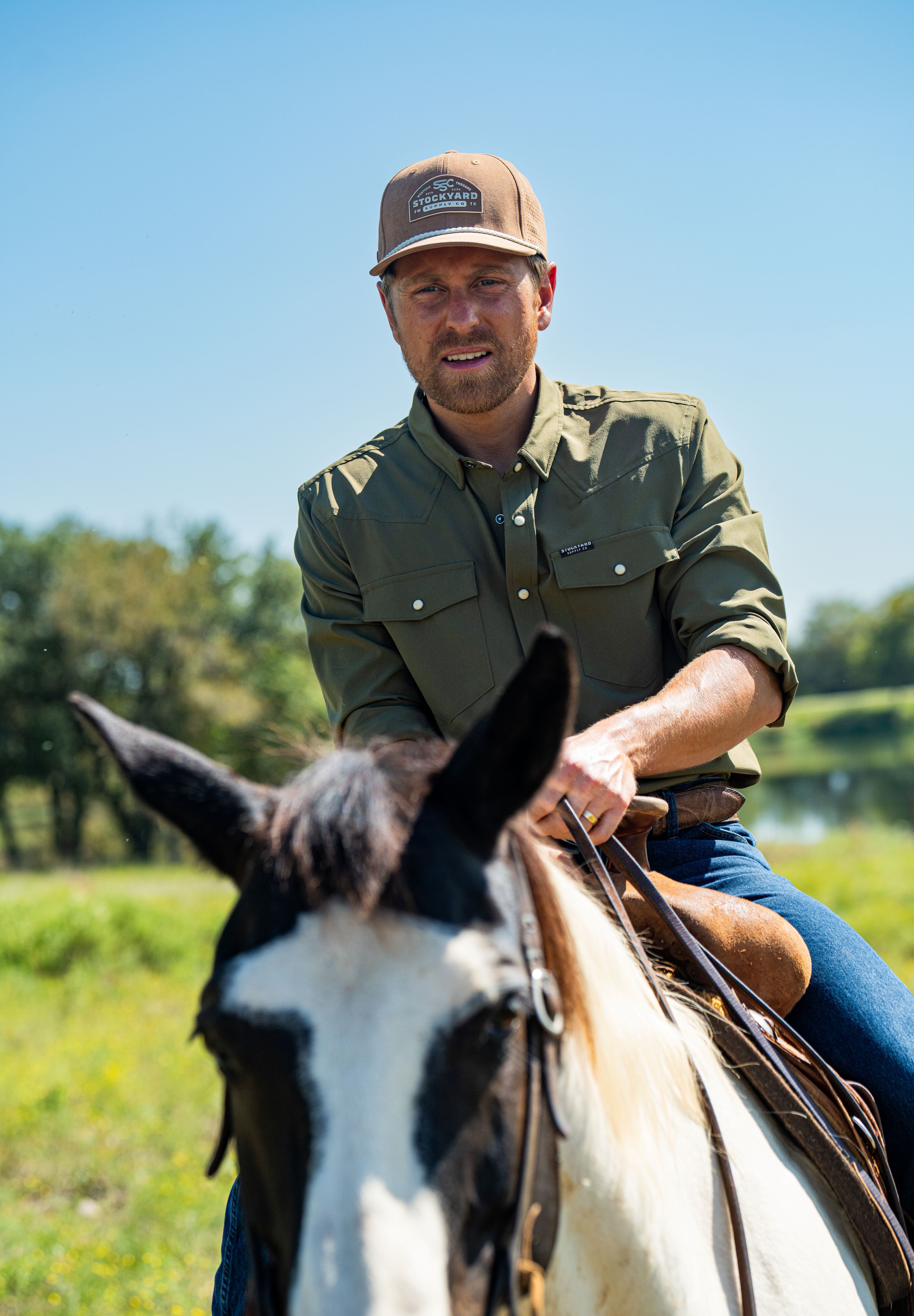 Eric Nelsen wearing the Coyote colored Champion hat and faded olive long sleeve performance pearl snap riding a horse.