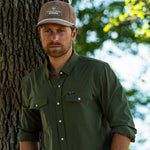 Eric Nelsen wearing the Coyote colored Champion hat and faded olive long sleeve performance pearl snap standing next to a tree. 