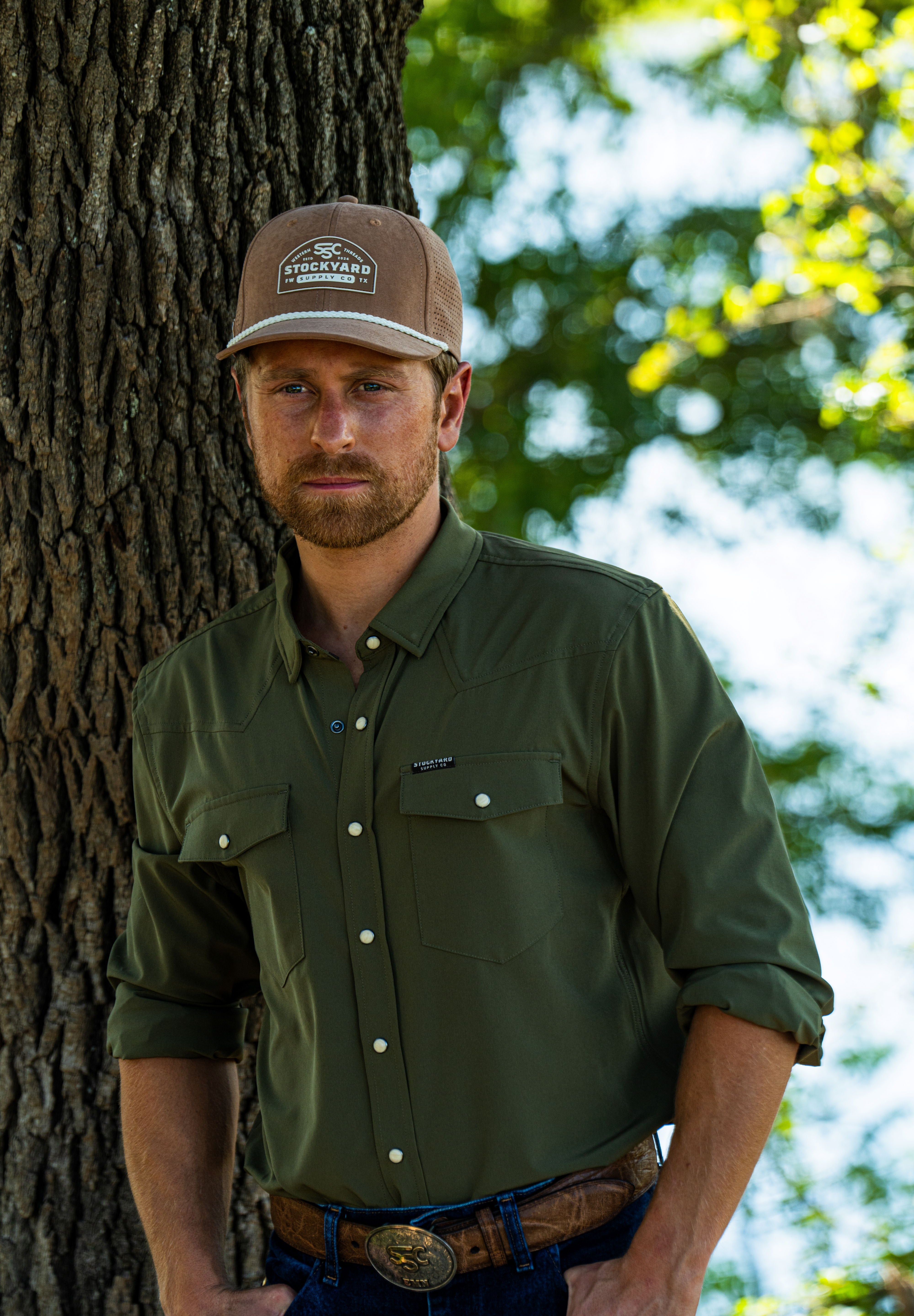 Eric Nelsen wearing the Coyote colored Champion hat and faded olive long sleeve performance pearl snap standing next to a tree. 