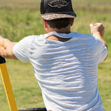 Eric Nelsen wearing a white shirt with the Commander Stockyard Supply Co hat with a black mesh and camo pattern brim unloading a wheelbarrow with his back to the camera. 