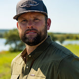 Ryan Dukes wearing a white shirt with the Commander Stockyard Supply Co hat with a black mesh and camo pattern brim and faded olive green pearl snap shirt. 