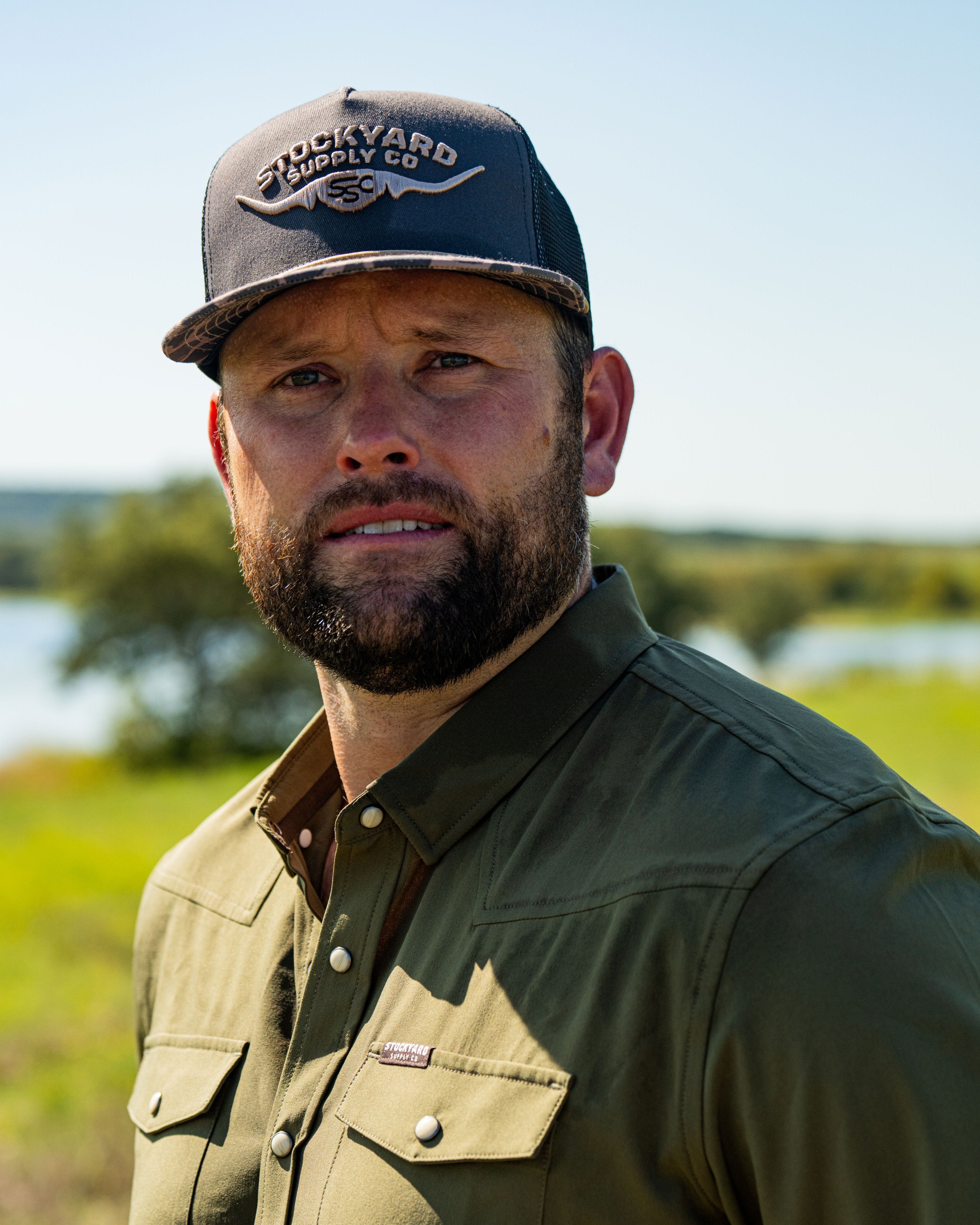 Ryan Dukes wearing a white shirt with the Commander Stockyard Supply Co hat with a black mesh and camo pattern brim and faded olive green pearl snap shirt. 