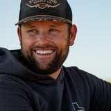 Ryan Dukes wearing a white shirt with the Commander Stockyard Supply Co hat with a black mesh and camo pattern brim with a Stockyard Supply Co hoodie leaning on a metal fence. 