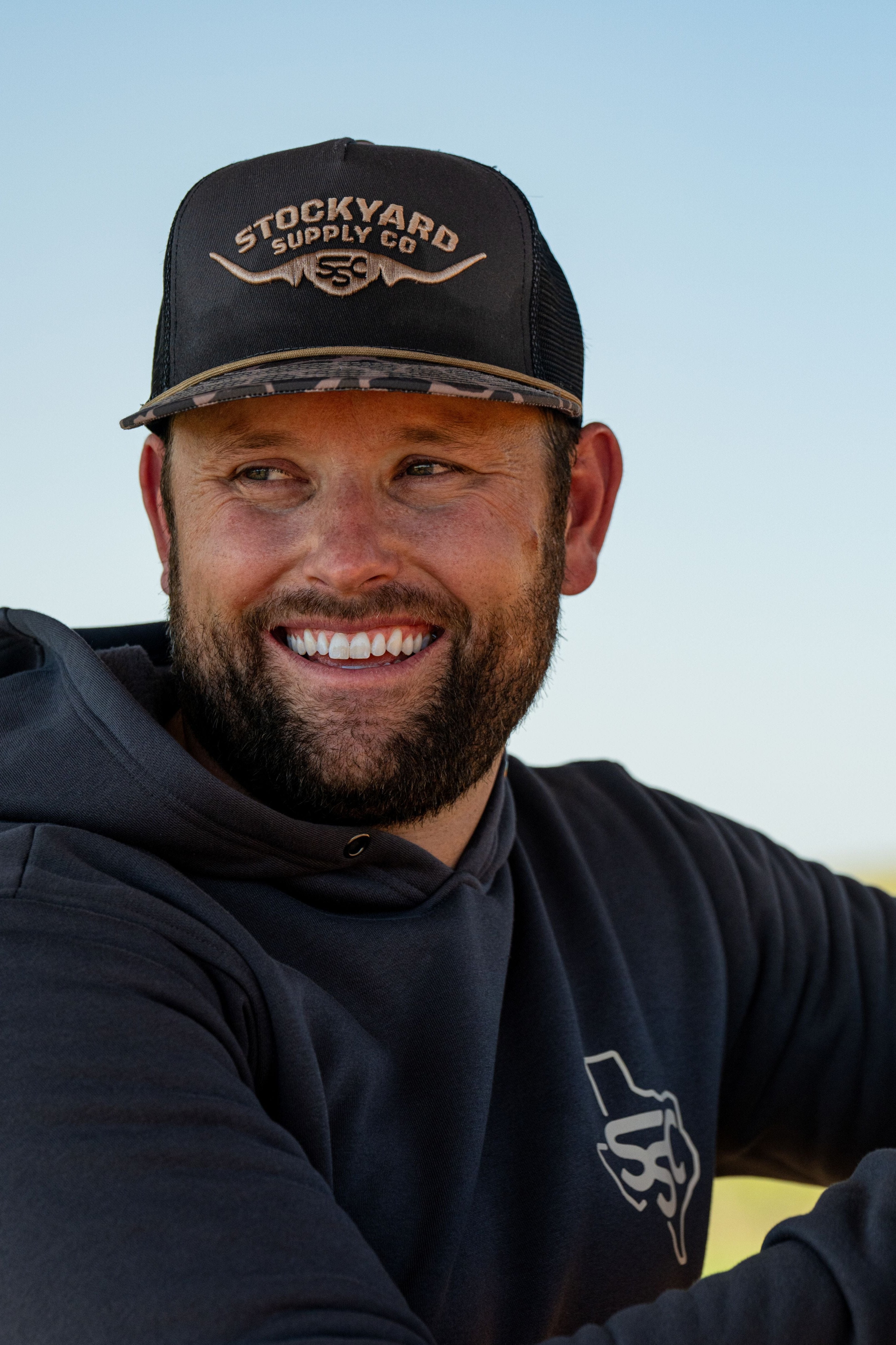 Ryan Dukes wearing a white shirt with the Commander Stockyard Supply Co hat with a black mesh and camo pattern brim with a Stockyard Supply Co hoodie leaning on a metal fence. 