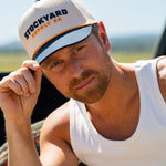 Eric Nelsen wearing a white tank top and the Two Step Stockyard Supply Co hat cream and navy colored. Holding the Navy bill. 