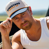 Eric Nelsen wearing a white tank top and the Two Step Stockyard Supply Co hat cream and navy colored. Holding the Navy bill. 