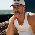 Eric Nelsen wearing a white tank top and the Two Step Stockyard Supply Co hat cream and navy colored sitting on a truck bed looking out on the horizon. 