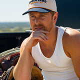 Eric Nelsen wearing a white tank top and the Two Step Stockyard Supply Co hat cream and navy colored sitting on a truck bed looking out on the horizon. 